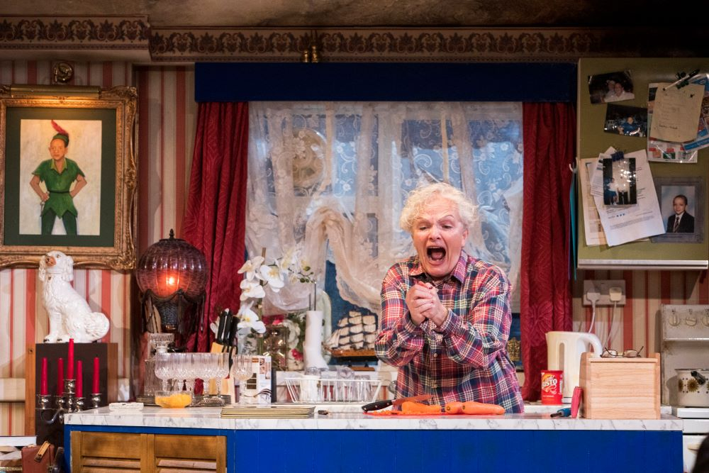 An older woman screaming with her hands clasped together. She has a pink checkered shirt on and is stood in a highly decorated kitchen.