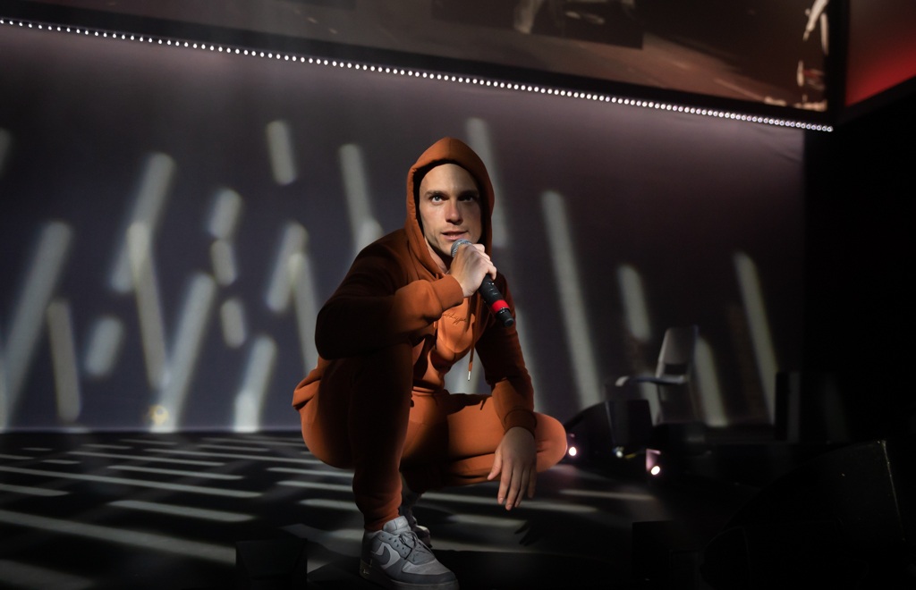 A man crouching with a burnt orange tracksuit. His hood is up and he is holding a microphone. The background has strips of white light on it