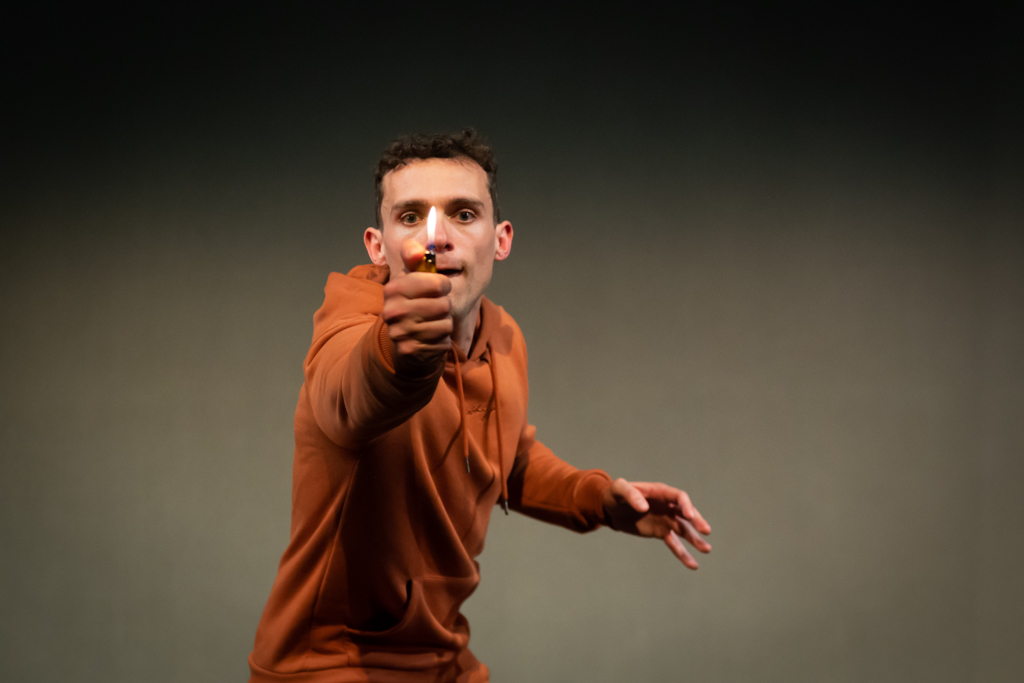 A man with short brown curly hair in a burnt orange tracksuit holding a lit lighter