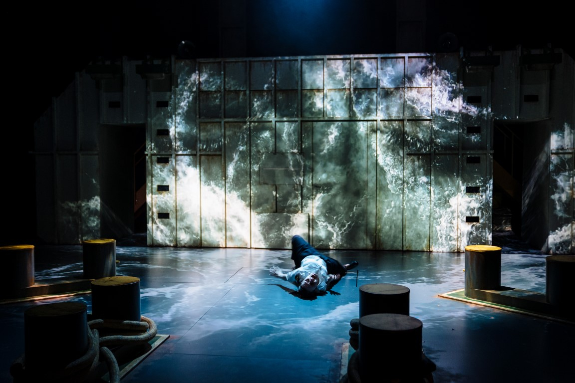 A man lying on the floor as if he fell into the ocean. There is a projection of waves on him and the shipping container.