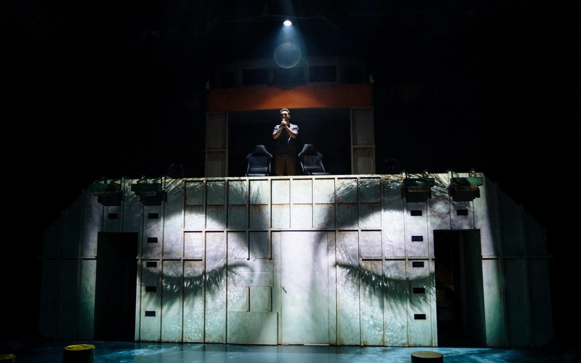 A pair of closed eyes projected onto a shipping container. A person is stood on top with a spotlight facing down on them as they have their hands in a prayer position.