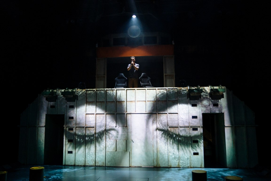 A pair of closed eyes projected onto a shipping container. A person is stood on top with a spotlight facing down on them as they have their hands in a prayer position.