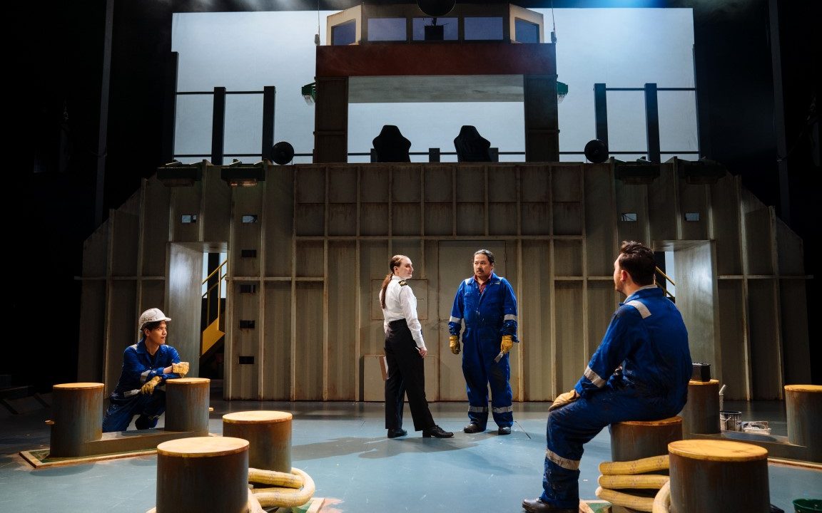 Three people wearing deep blue boiler suits. The set has six cleats and is set up like a cargo ship. One is sat on Cleat, whilst another is tieing rope to one. The last person is stood talking to a person wearing a boat staff uniform.