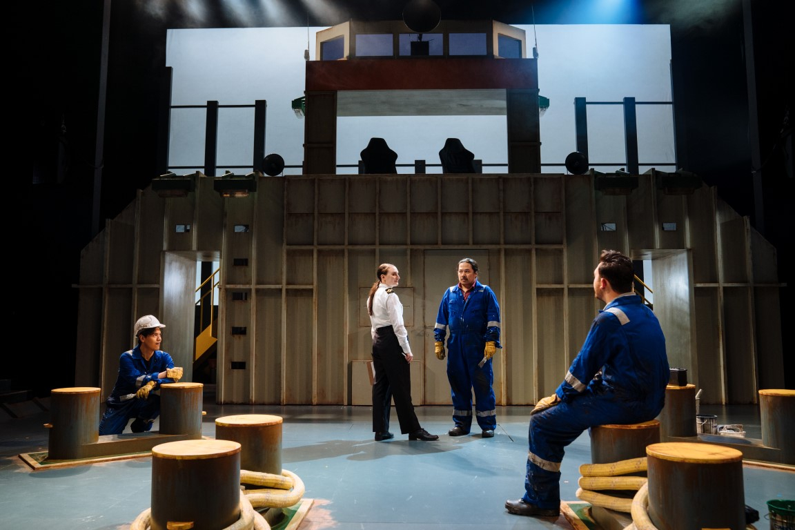 Three people wearing deep blue boiler suits. The set has six cleats and is set up like a cargo ship. One is sat on Cleat, whilst another is tieing rope to one. The last person is stood talking to a person wearing a boat staff uniform.