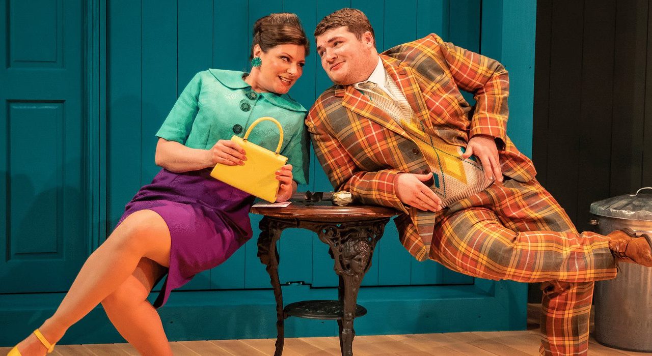 A man and woman leaning on a coffee table. The woman is wearing a button up turquoise top, purple skirt and yellow heels which match her bag which she is holding with two hands. The man has an orange checkered suit on.