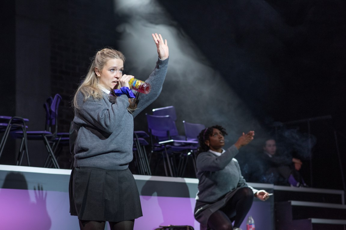 A blonde girl in a grey school uniform is stood with one hand raised and the other holding a microphone. Behind her their is another girl in a grey school oufit crouched down with her arm out.