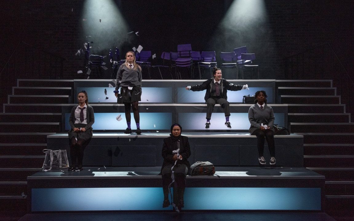 Five school girls dressed in grey school uniforms. Two of them are stood on a stair case of four light up blocks, the others are sat on them.