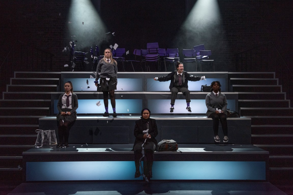 Five school girls dressed in grey school uniforms. Two of them are stood on a stair case of four light up blocks, the others are sat on them.