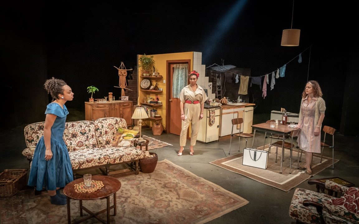 Three women in a living room stood speaking across to eachother. The Living room is well furnished and warm with a 50's style.