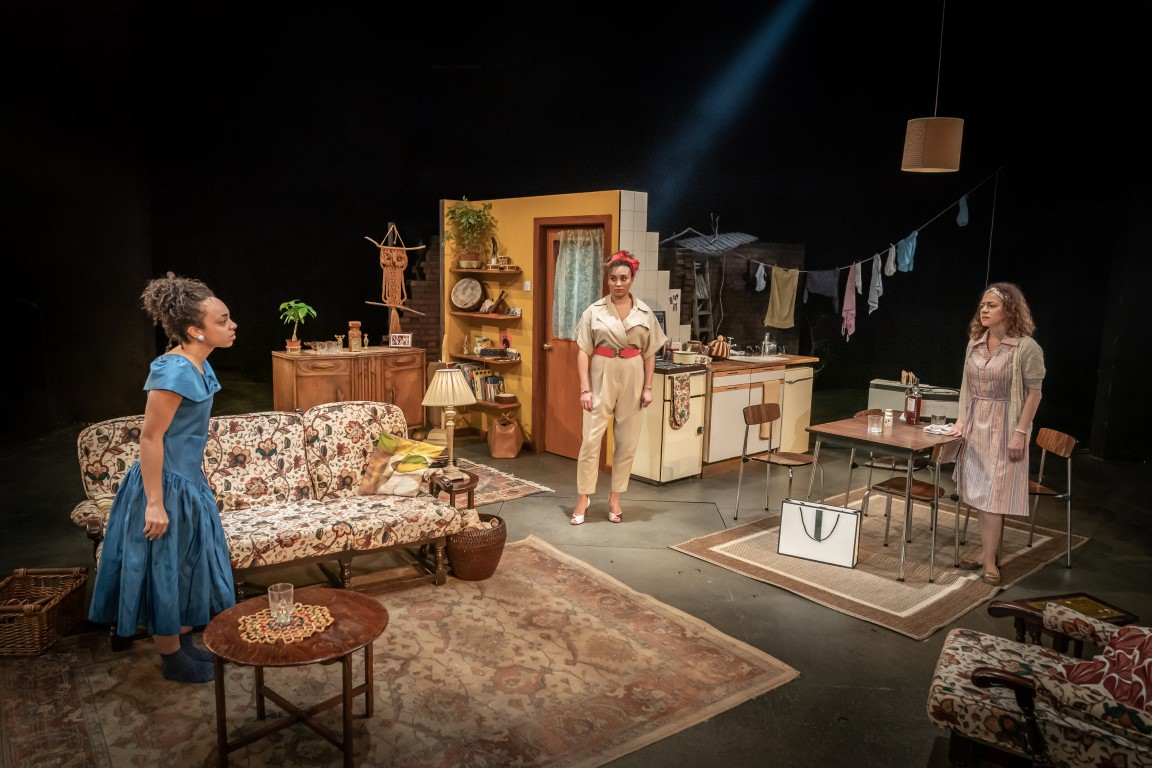 Three women in a living room stood speaking across to eachother. The Living room is well furnished and warm with a 50's style.