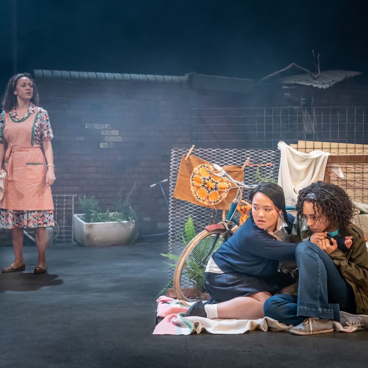 Two women crouched behind a bike and some washing hiding from a woman who is stood behind them.