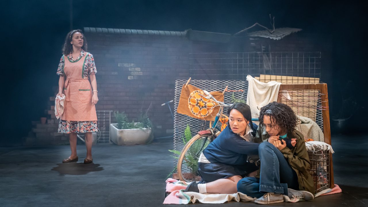 Two women crouched behind a bike and some washing hiding from a woman who is stood behind them.