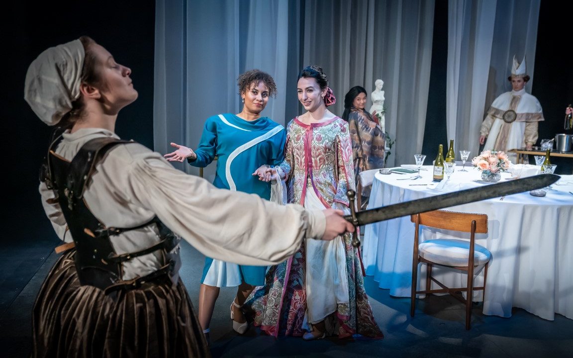 A woman wearing a medieval dress and bonnet holding a sword. Two are stood across from her staring. In the background there is a dinner party and guests.