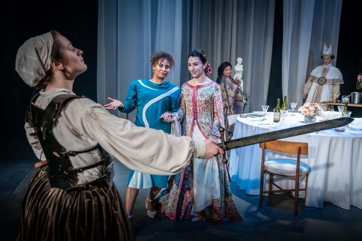 A woman wearing a medieval dress and bonnet holding a sword. Two are stood across from her staring. In the background there is a dinner party and guests.