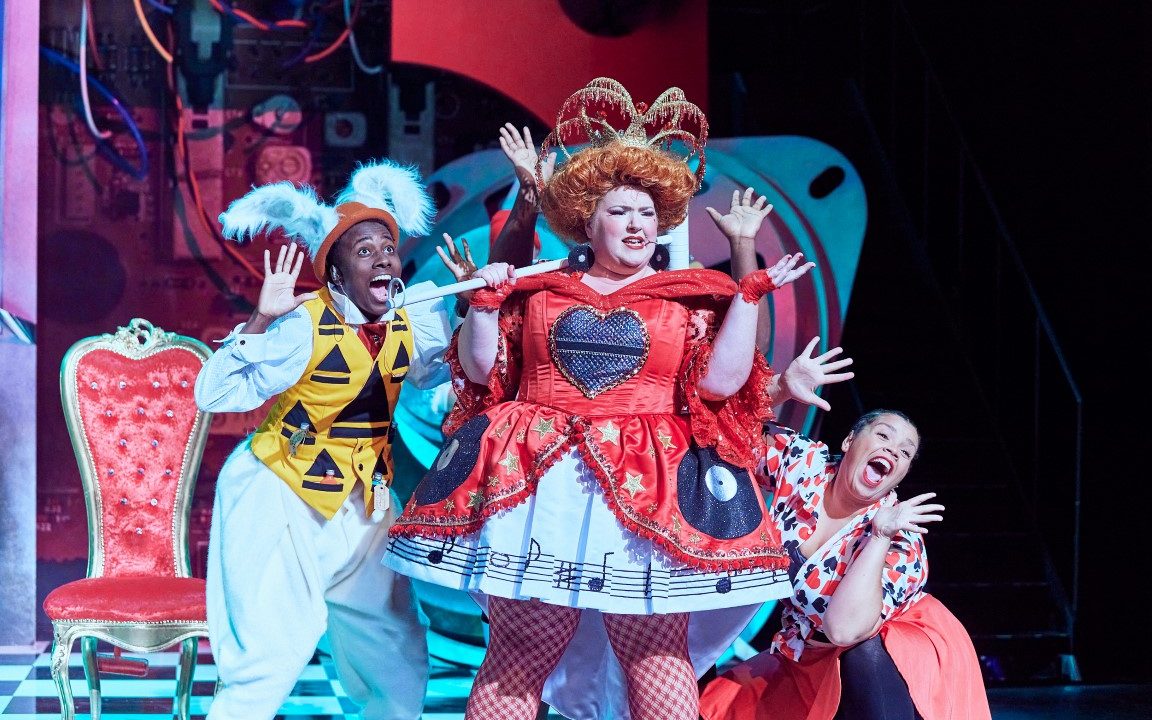 A woman with a crown, ginger wig and red velvet dress is singing into her scepter. Behind her are to other cast members also singing