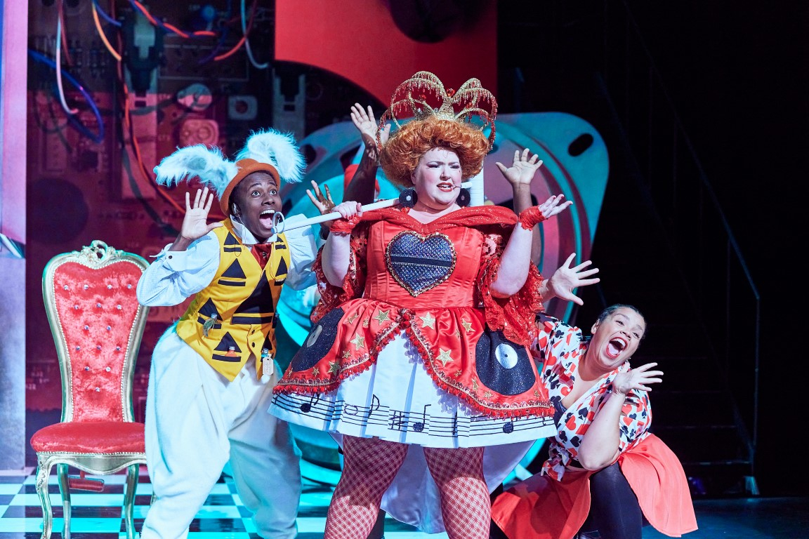 A woman with a crown, ginger wig and red velvet dress is singing into her scepter. Behind her are to other cast members also singing