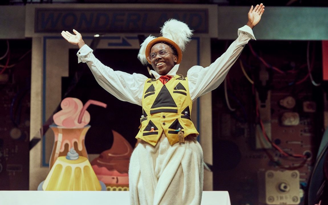 A man with rabbit ears, a yellow vest and a large pocket watch has his arms spread and is smiling