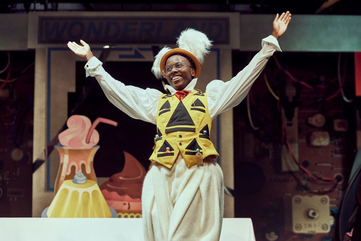 A man with rabbit ears, a yellow vest and a large pocket watch has his arms spread and is smiling