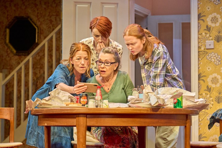 All cast members gathered around a phone. Two are sat at the table which is covered in the aftermath of a chippy. They all look concerned.