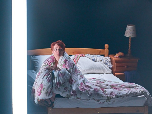 A woman with short hair sat on the edge of the bed with her cover cocooning around her.