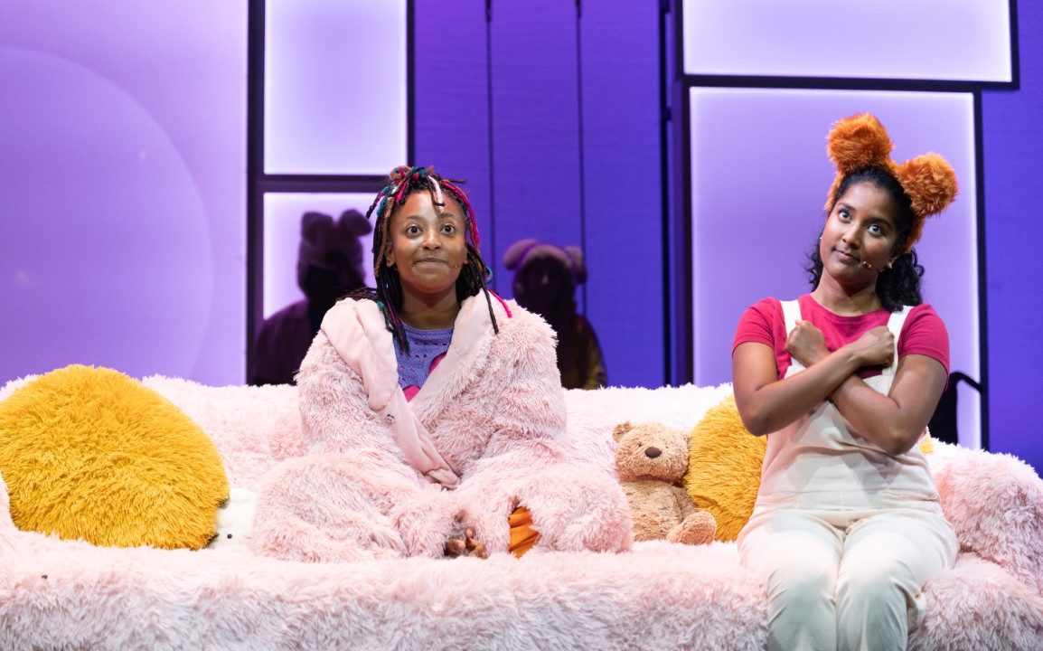 two women sat on a fluffy pink sofa. One is wearing a pink fluffy dressing gown and the other a pink top and dugarees. The woman with dungarees also has a brown bear ear headband on.