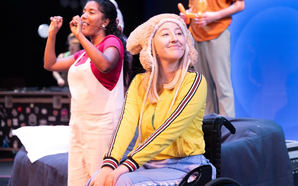 A woman wearing a brown fluffy hat, yellow jumper and purple trousers is sat smiling in a wheelchair. In the background is another woman wearing a pink bow, top and dungarees. She is stood and signing