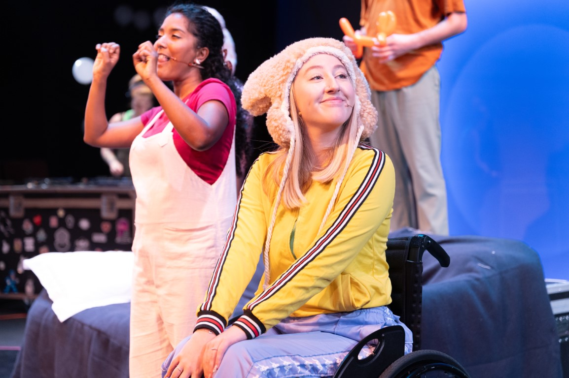 A woman wearing a brown fluffy hat, yellow jumper and purple trousers is sat smiling in a wheelchair. In the background is another woman wearing a pink bow, top and dungarees. She is stood and signing