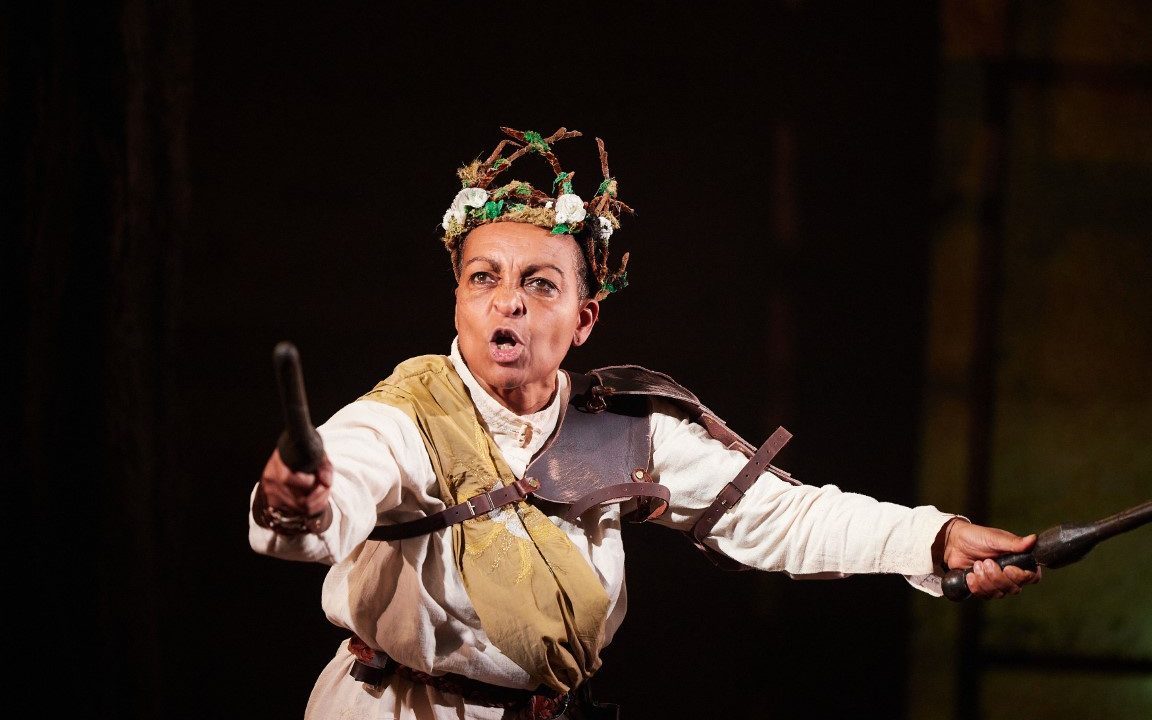 Richard the third wearing a branch head piece, a white shirt, leather armor and a beige sash. They are holding a brown baton in each hand.