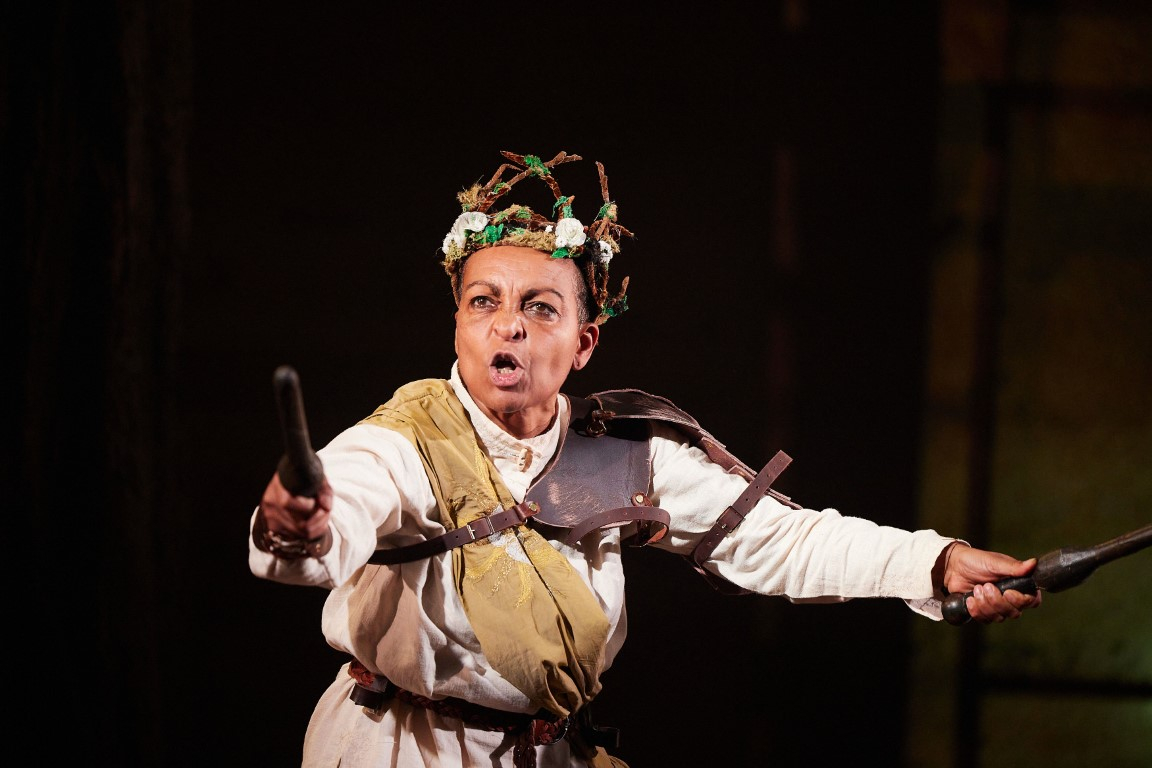 Richard the third wearing a branch head piece, a white shirt, leather armor and a beige sash. They are holding a brown baton in each hand.