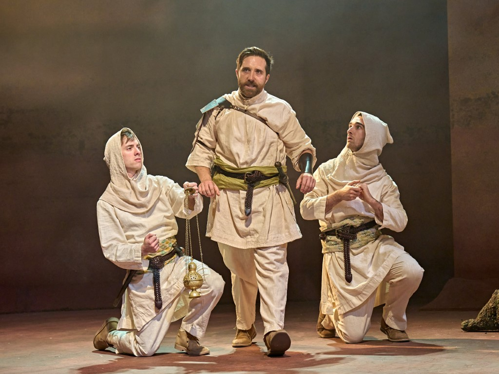 A man walking forward, wearing a beige baggy shirt and trousers. Behind him are to people who are hooded and kneeling.