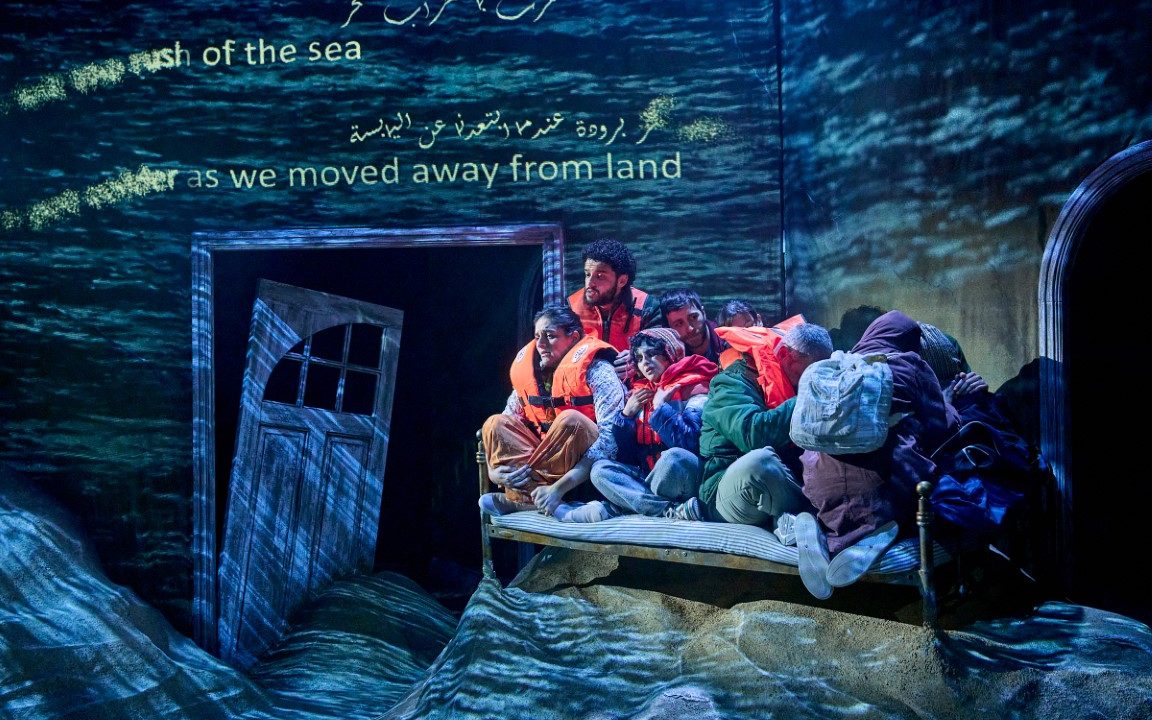 the Cast all sat on a single bed as if they were on a small raft wearing life jackets. The set has an ocean blue wash to make it appear they are at sea. The floor is covered in sand.