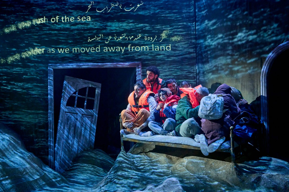 the Cast all sat on a single bed as if they were on a small raft wearing life jackets. The set has an ocean blue wash to make it appear they are at sea. The floor is covered in sand.