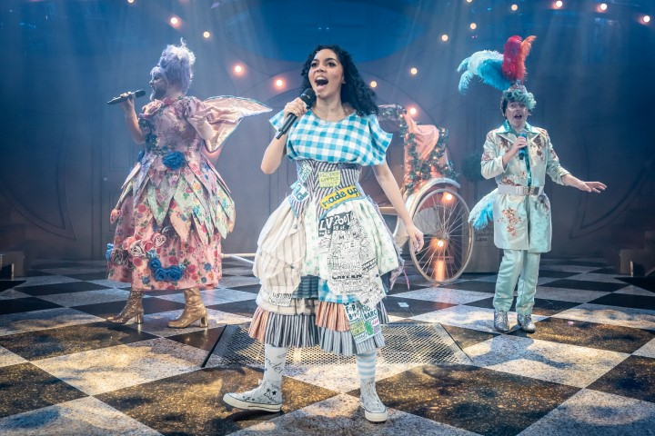 Cinderella singing into a microphone. She is wearing a checkered blue top, white baggy trousers with tassels and white boots. Behind her are two other cast members also singing.