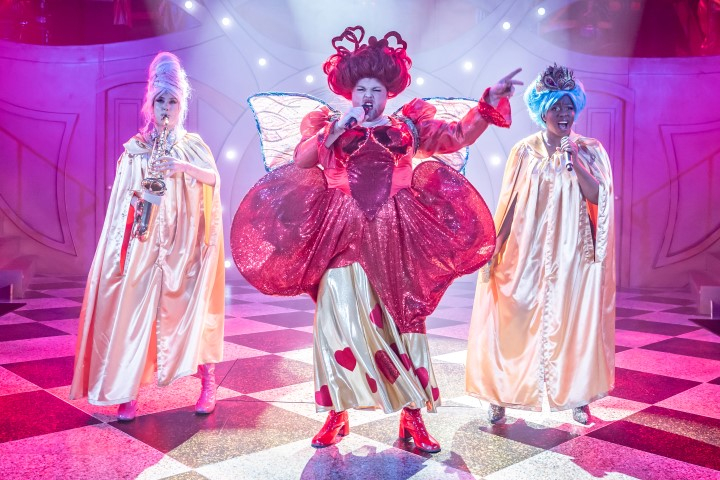 The Fairy God mother stood singing into a microphone. They are wearing a big red wig, red and gold butterfly wings, a gold skirt with love hearts on and red healed boots. Behind them are two cast members, one is also singing into a microphone the other is playing the saxophone.