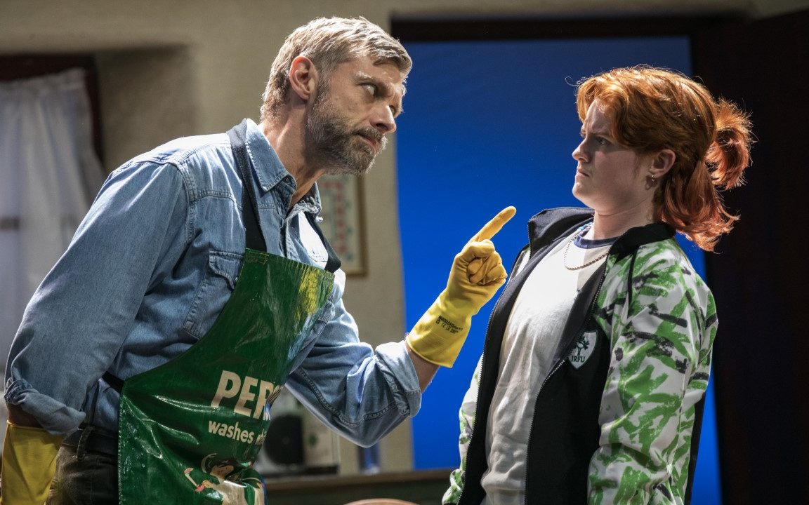 Two people stood in a kitchen. The person on the left has grey hair and is wearing a blue shirt covered by a green apron and yellow rubber gloves. They are pointing at the person on the right. The person on the right has ginger hair tied up and is wearing a grey top, chain and green zip up jacket.