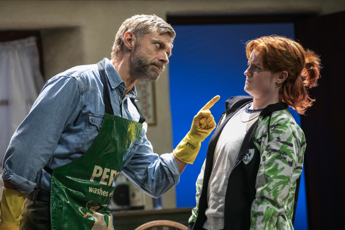 Two people stood in a kitchen. The person on the left has grey hair and is wearing a blue shirt covered by a green apron and yellow rubber gloves. They are pointing at the person on the right. The person on the right has ginger hair tied up and is wearing a grey top, chain and green zip up jacket.