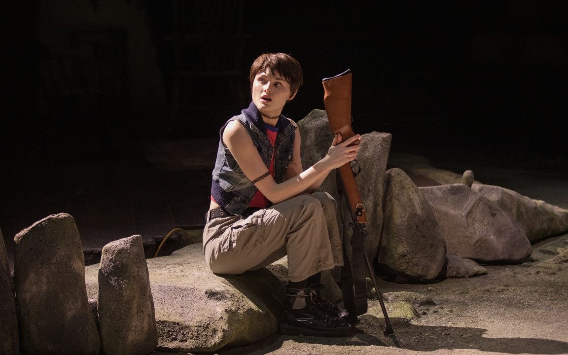 A person with short brown hair sat on a rock holding a rifle gun. They are wearing a vest, long trousers and chunky black boots.