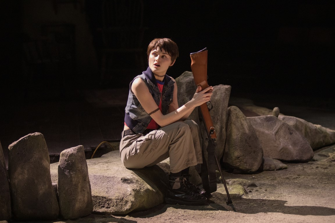 A person with short brown hair sat on a rock holding a rifle gun. They are wearing a vest, long trousers and chunky black boots.