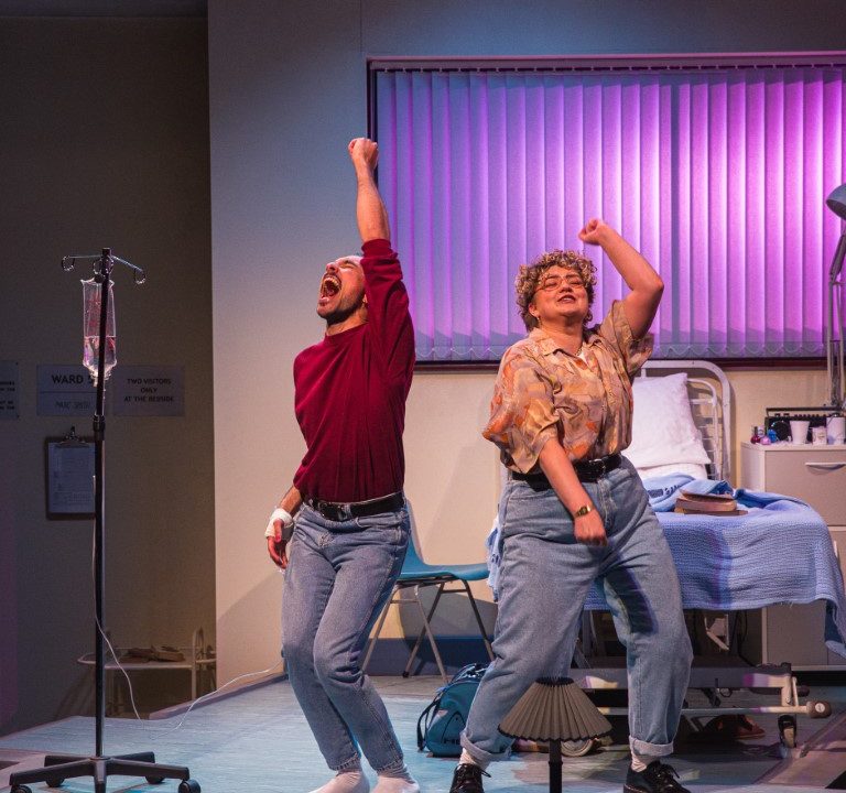 Two people dancing in a hospital room with their arms in the air.