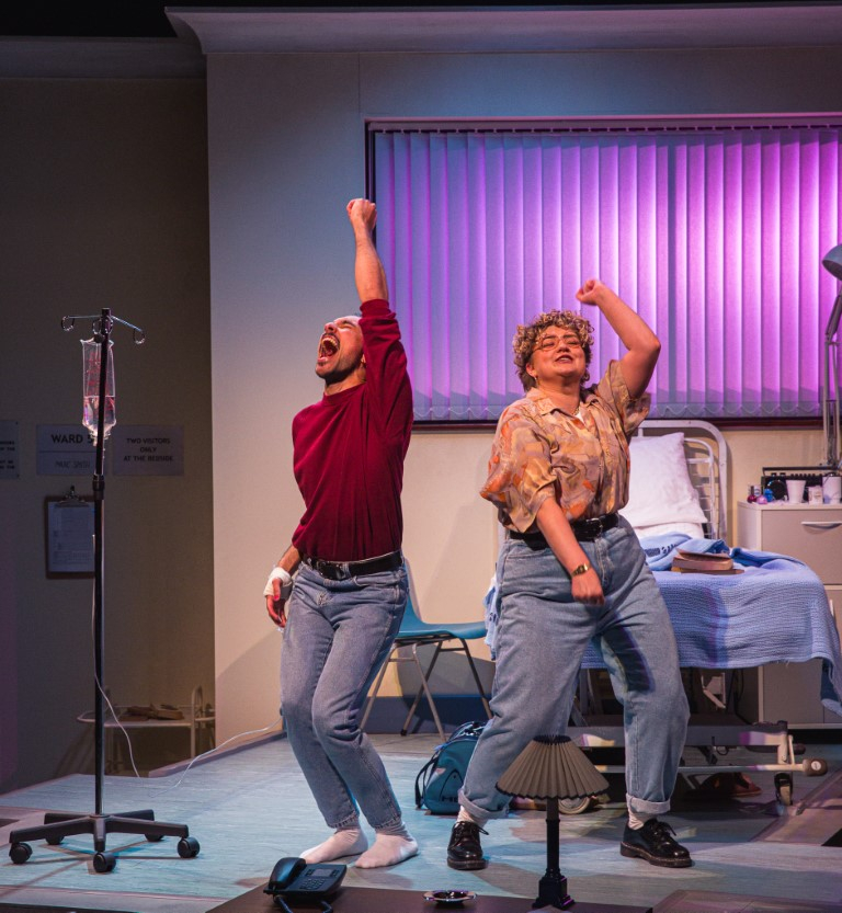 Two people dancing in a hospital room with their arms in the air.