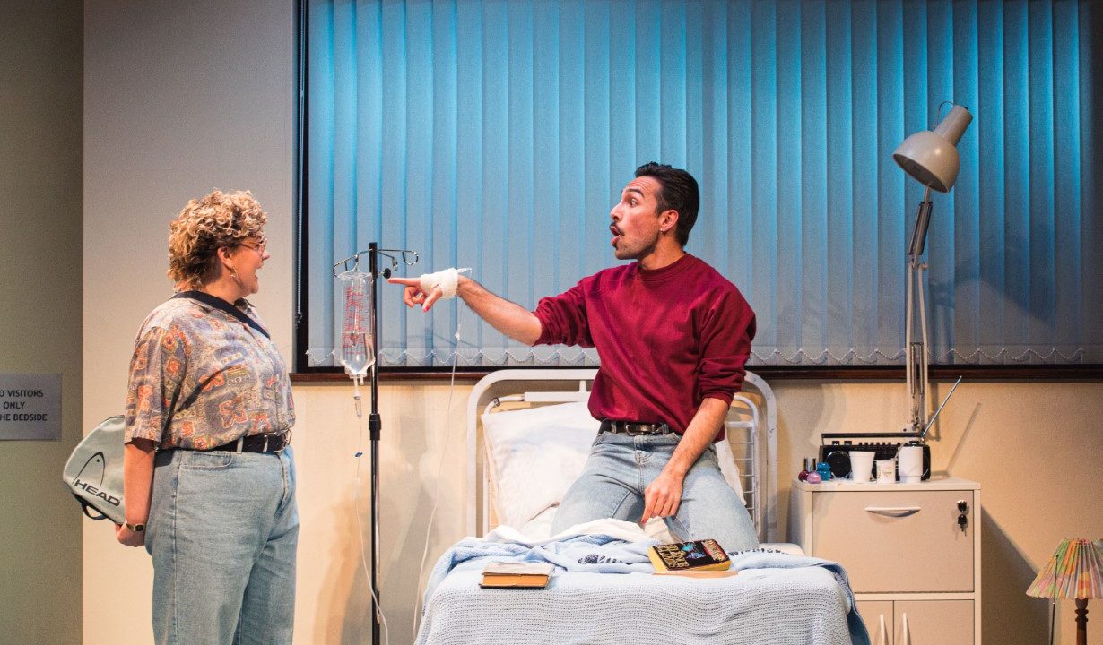 Two people in a hospital room. On the hospital bed is a man kneeling and pointing at a woman