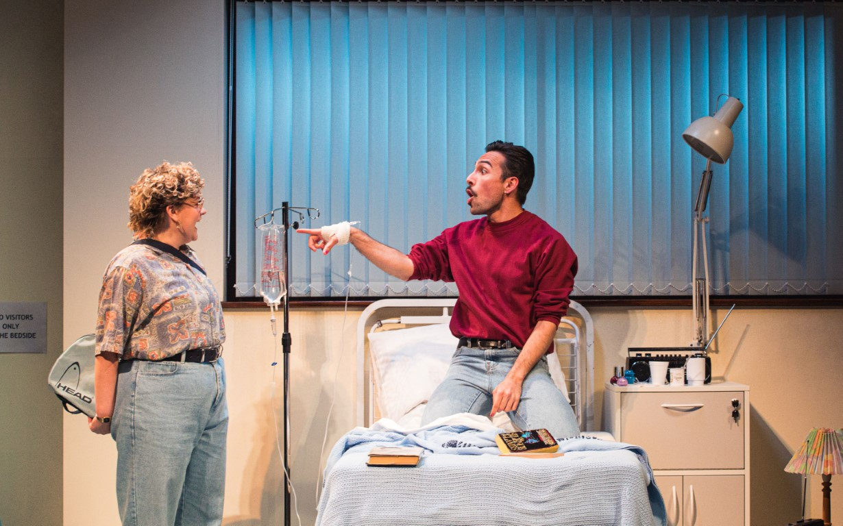 Two people in a hospital room. On the hospital bed is a man kneeling and pointing at a woman