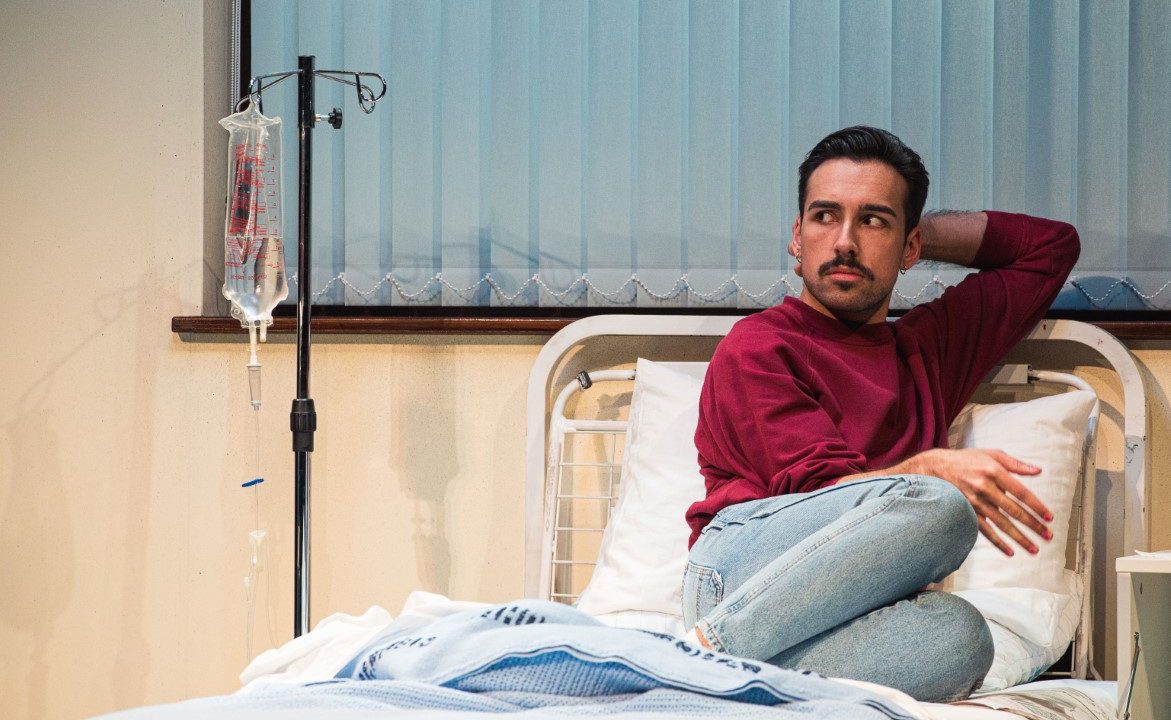 A man sat with short dark hair and a moustache is sat with his knees up on the top of a hospital bed. Next to him is an IV drip bag and stand. He is wearing a red long sleeve top and light wash blue jeans.