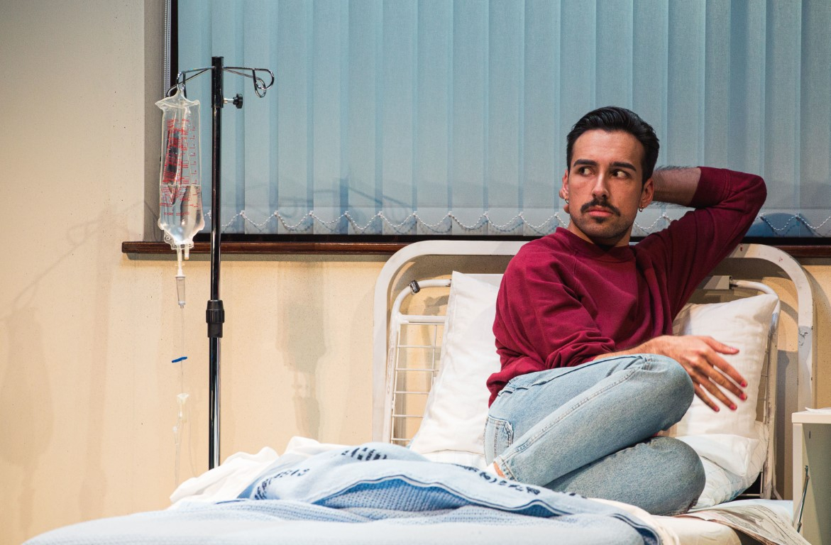 A man sat with short dark hair and a moustache is sat with his knees up on the top of a hospital bed. Next to him is an IV drip bag and stand. He is wearing a red long sleeve top and light wash blue jeans.