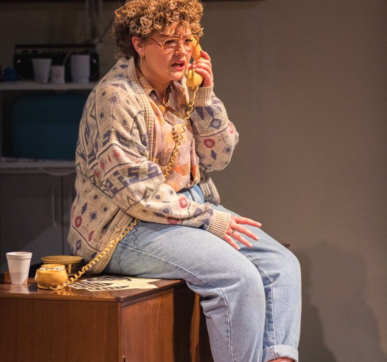 a woman with short curly blonde hair is wearing large glasses and is sat on a cabinet. She is also wearing a knitted cardigan, shirt , jeans and slippers. On the cabinet is a yellow corded phone which she is holding up to her ear.