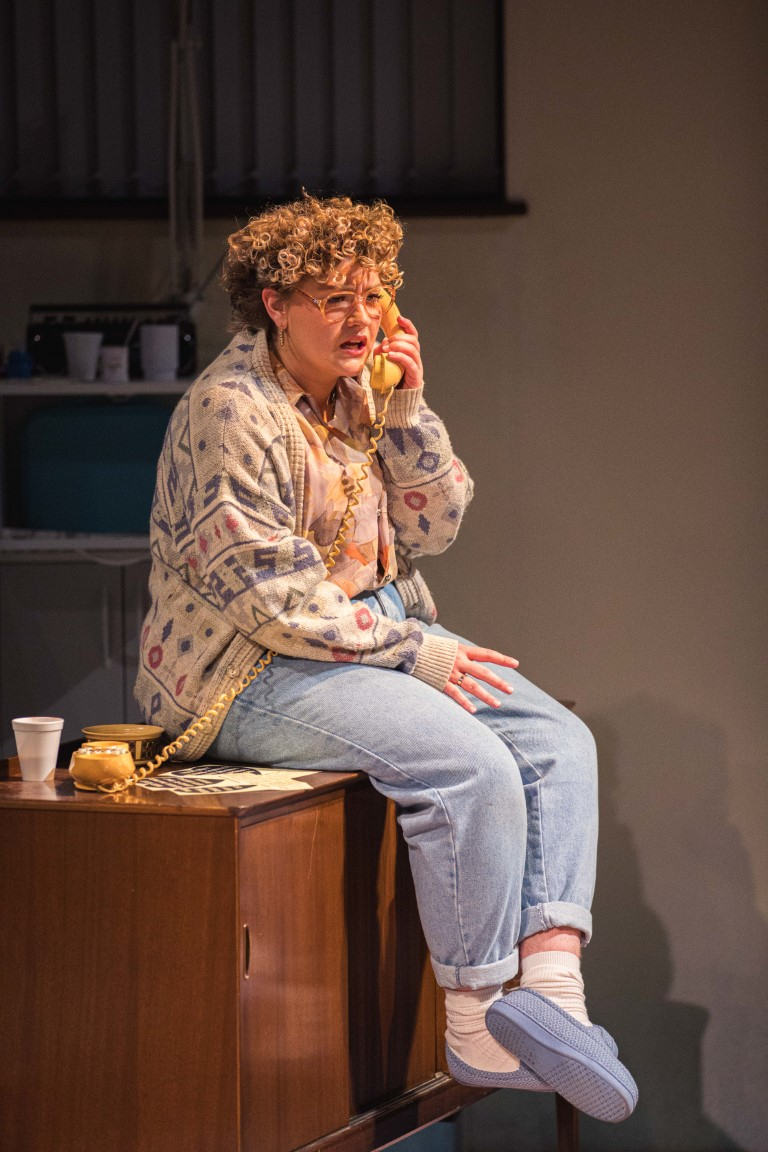 a woman with short curly blonde hair is wearing large glasses and is sat on a cabinet. She is also wearing a knitted cardigan, shirt , jeans and slippers. On the cabinet is a yellow corded phone which she is holding up to her ear.