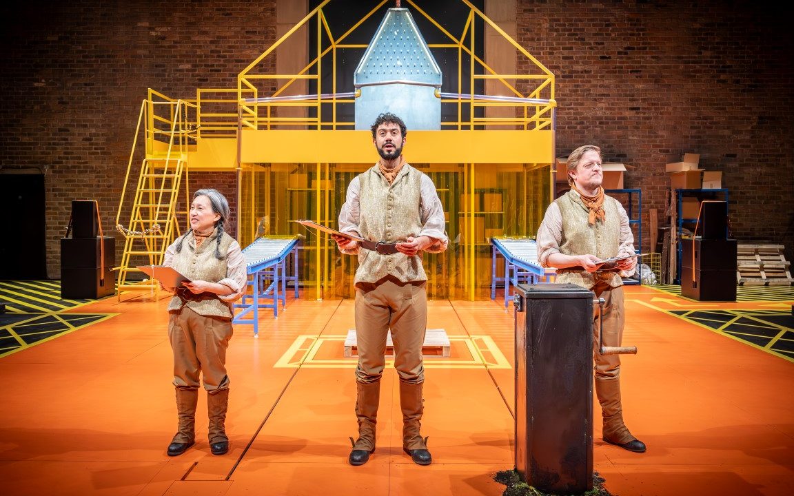 Three people stood holding clipboards. In the Back ground there is a Yellow pipe house with a set of stairs leading to a second floor.