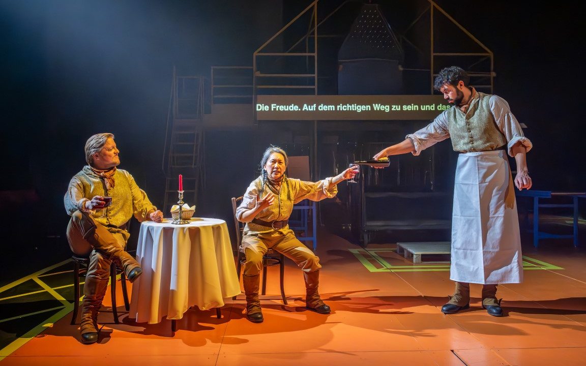 two people cat on chairs around a table with a white cloth, candle and bread basket on top of it. There is a third person wearing an apron serving them.