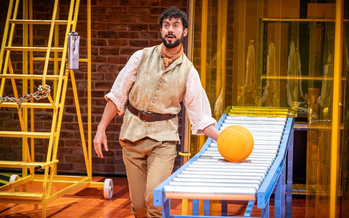 A tall man with brown curly hair, a moustache and beard. He is wearing a white floaty blouse, beige vest and trousers and an orange neckerchief. He is holding a ball that is resting on a conveyer belt.