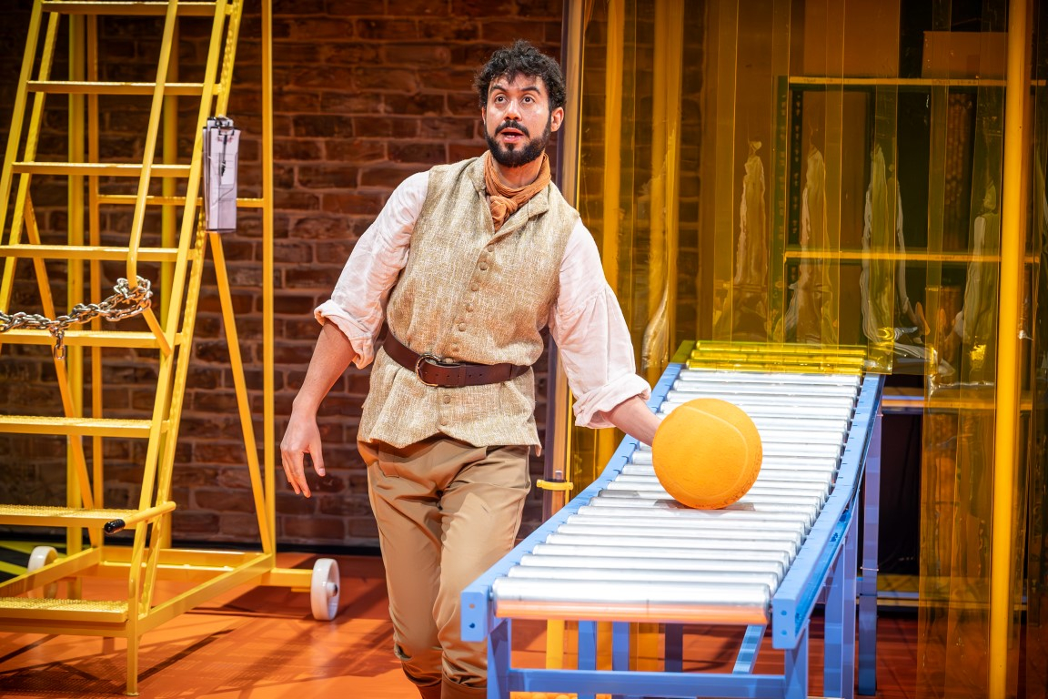 A tall man with brown curly hair, a moustache and beard. He is wearing a white floaty blouse, beige vest and trousers and an orange neckerchief. He is holding a ball that is resting on a conveyer belt.